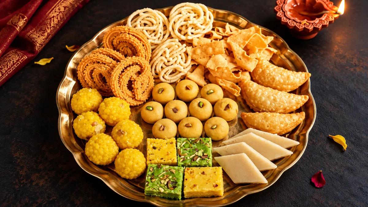 A brass platter of Indian Diwali sweets and savory snacks, including laddus, spiral snacks, barfi, and fried pastries, beside a lit diya.